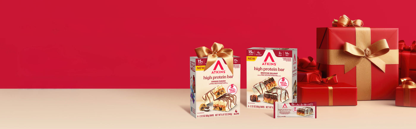 Boxes of Atkins high protein bars and wrapped gifts with gold bows are on a beige surface against a red background, creating a festive holiday feel.