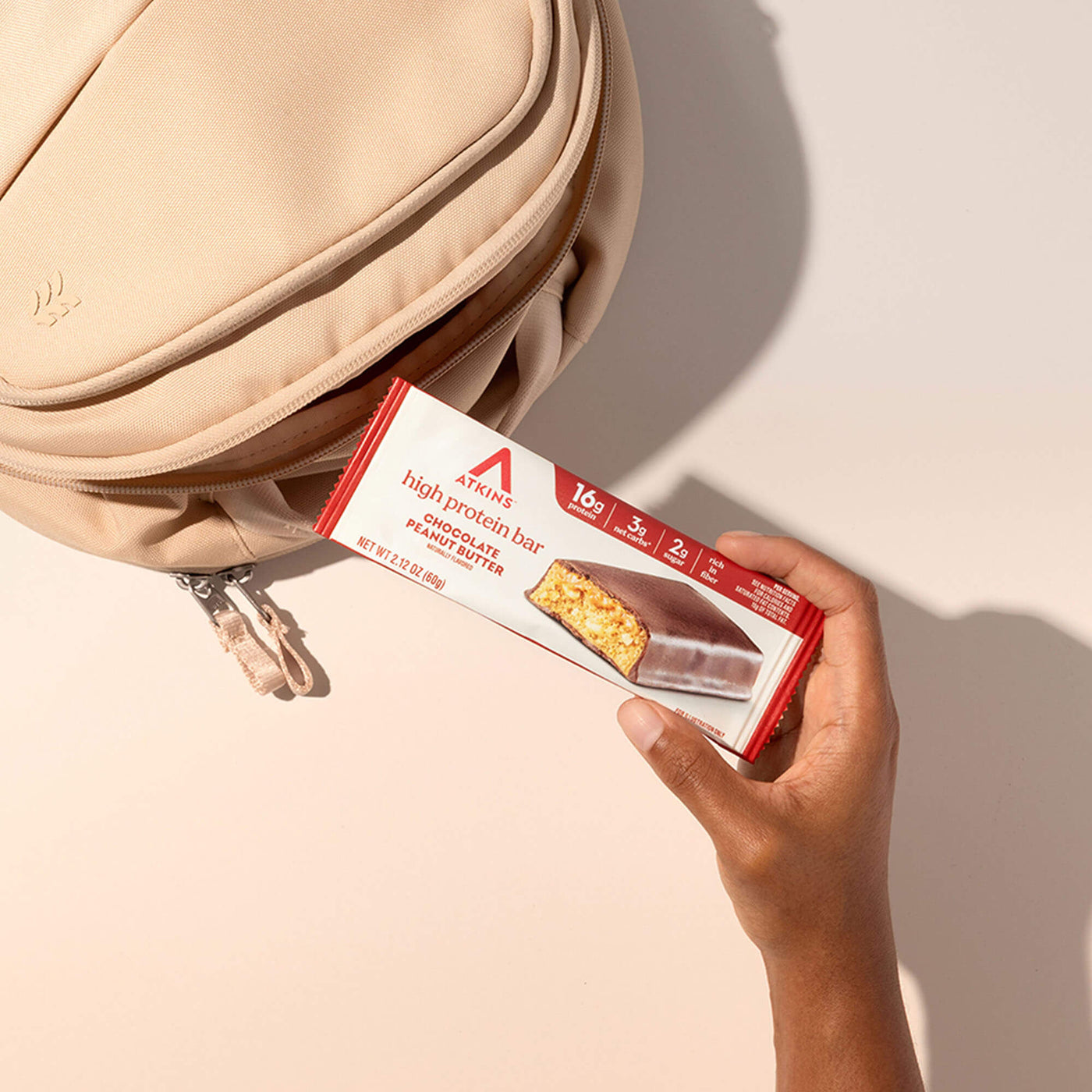 Hand holding a Atkins high protein bar package next to a beige bag on a light background