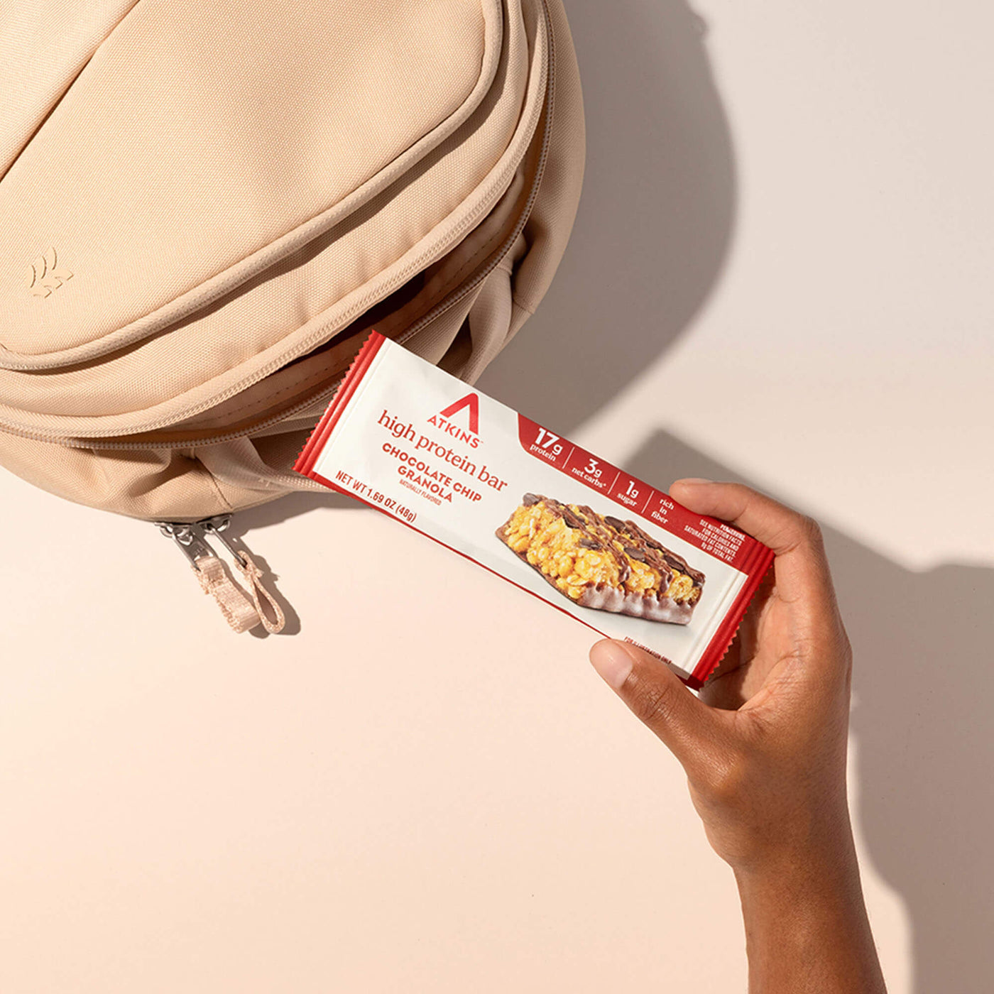 Hand holding a Atkins high protein bar package next to a beige backpack on a light background