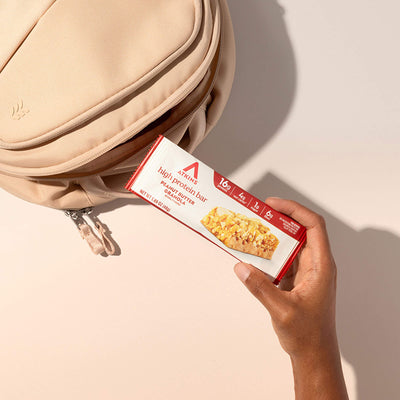 Hand holding a protein bar box in front of a beige bag on a light background