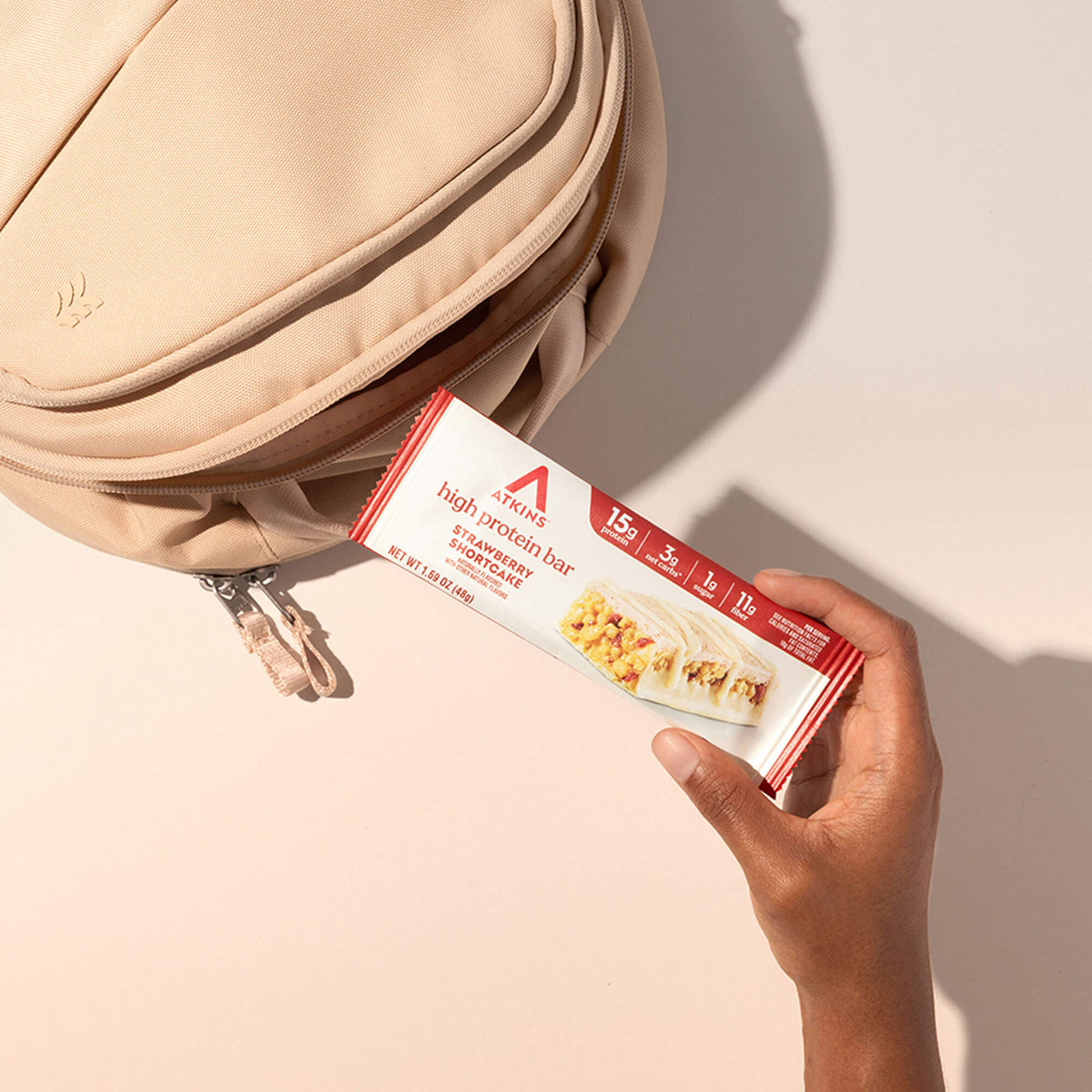 Hand holding Atkins high protein bar package next to a beige bag on a light background