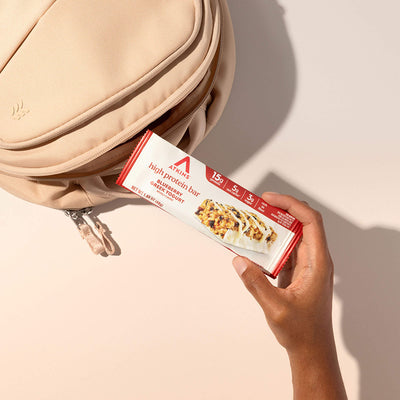 Hand holding a protein bar box in front of a beige bag on a light background