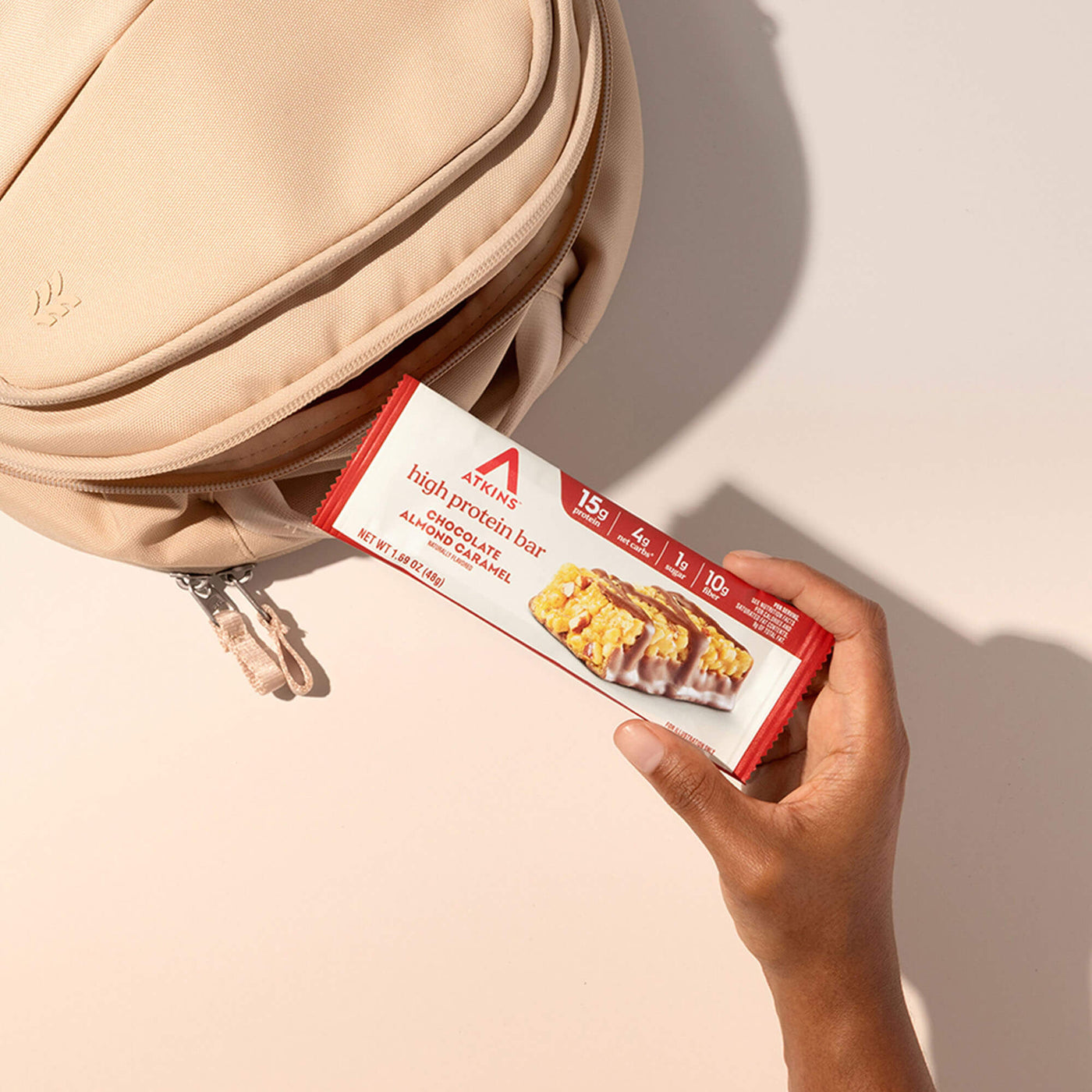 Hand holding a protein bar package in front of an open beige bag on a light background